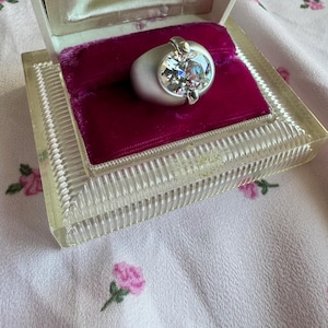 May include: A silver ring with a large, clear, round gemstone, displayed in a clear plastic box with a magenta velvet interior. The ring has a wide band and is set with two silver accents. The box is on a pink floral fabric.