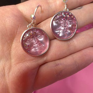 May include: A pair of round drop earrings featuring a translucent pink glass cabochon with a molded sun face design. The circular face is accented with small, iridescent multicolored crystals along the upper edge, set in a silver-toned metal frame.