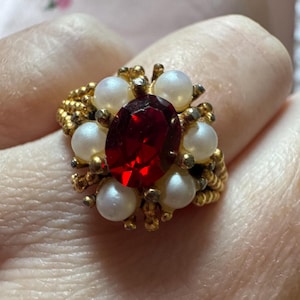 May include: A gold-toned ring featuring a large, oval, red gemstone at the center, surrounded by smaller, white, pearl-like accents. The ring's band has a textured, braided design, creating a vintage or antique aesthetic.