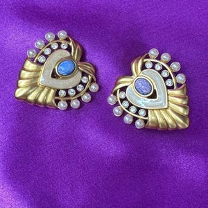 Op de afbeelding: Een paar hartvormige oorbellen met een afwerking van geborsteld goudkleurig metaal, een centrale ovale blauwe cabochon, crèmekleurige emaille accenten en een rand versierd met kleine witte kunstparels en heldere ronde strass-steentjes.