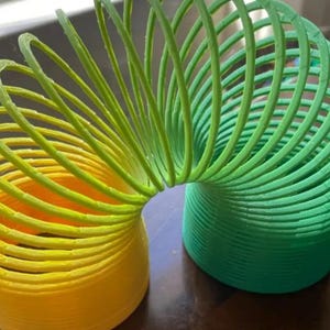 Puede incluir: Un juguete slinky multicolor en forma de espiral. El juguete pasa del amarillo al verde, con la sección verde en el lado derecho. El juguete es de plástico y está sobre una superficie marrón.