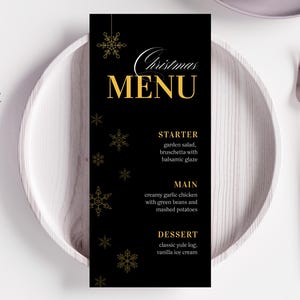 Może przedstawiać: Czarna karta menu bożonarodzeniowego ze złotym tekstem i akcentami płatków śniegu, umieszczona na białym talerzu ze sztućcami. Menu zawiera opcje przystawek, dań głównych i deserów. Świąteczny motyw.