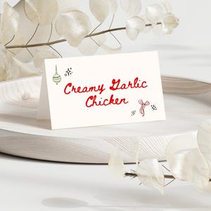 May include: A folded white card with the text "Creamy Garlic Chicken" in red script, with decorative elements. The card sits on a light wooden tray, with white foliage in the background.