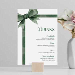May include: A white drinks menu with a green watercolor bow and ribbon design. The menu lists cocktails, wine, beer, and non-alcoholic beverages. A white vase with flowers is to the right.