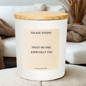 May include: A white cylindrical candle with a wooden lid. The candle has a label with the text "SOLACE STUDIO" and the phrase "TRUST NO ONE. ESPECIALLY YOU." It is a soy wax candle, 9oz.