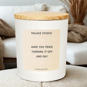 May include: A white soy wax candle with a wooden lid. The label reads "SOLACE STUDIO" and "HAVE YOU TRIED TURNING IT OFF AND ON?". The candle is 90 oz.