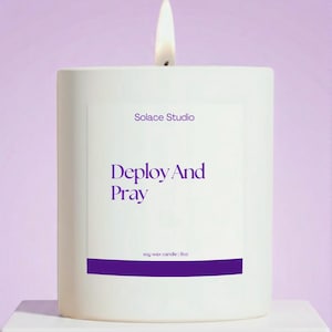 May include: A lit white soy wax candle with the text "Deploy And Pray" in purple. The candle is from Solace Studio and is 9oz. The background is a soft purple.