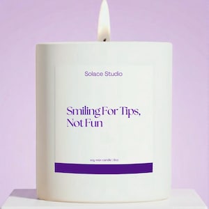 May include: A white rectangular candle with a lit wick, set against a soft purple background. The candle has the text "Solace Studio" and "Smiling For Tips, Not Fun" in purple. It is labeled as a 9oz soy wax candle.