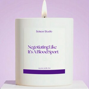 May include: A white soy wax candle with a lit wick. The candle has a purple label with the text "Negotiating Like It's A Blood Sport" and "Solace Studio" printed on it. The candle is 9oz.