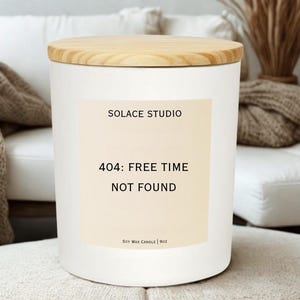 May include: A white soy wax candle with a wooden lid. The candle has a label that reads "SOLACE STUDIO" and "404: FREE TIME NOT FOUND". The candle is 9oz.
