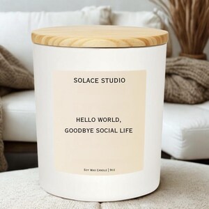 May include: A white soy wax candle with a wooden lid. The candle has a label that reads "SOLACE STUDIO" and "HELLO WORLD, GOODBYE SOCIAL LIFE." The candle is 9oz.