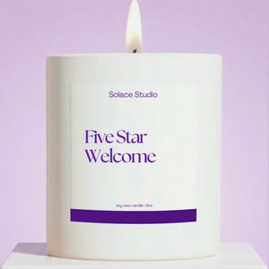 May include: A lit white soy wax candle with the text "Solace Studio" and "Five Star Welcome" in purple. The candle is 9oz and sits on a white surface against a lavender background.