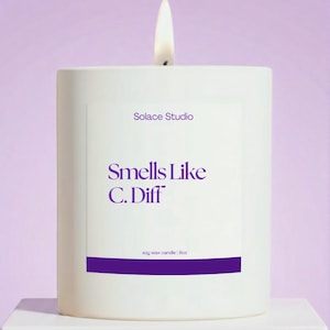 May include: A lit white soy wax candle with the text "Solace Studio" and "Smells Like C. Diff" in purple. The candle is 9oz and sits on a white surface against a purple background.
