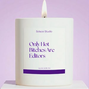 May include: A white soy wax candle with a lit wick. The candle has the text "Solace Studio" and "Only Hot Bitches Are Editors" in purple. The candle is 9oz.
