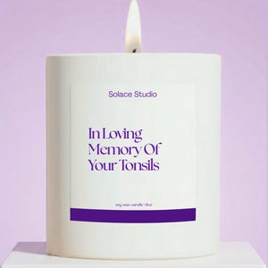 May include: A lit white soy wax candle with the text "In Loving Memory Of Your Tonsils" in purple. The candle is from Solace Studio and is 9oz. The background is a soft purple.