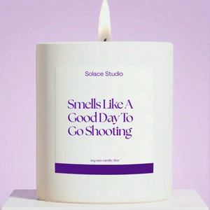 May include: A lit white soy wax candle with the text "Smells Like A Good Day To Go Shooting" in purple. The candle is from Solace Studio and is 9oz. The background is a soft purple.