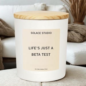 May include: A white soy wax candle with a wooden lid. The candle has a label that reads "SOLACE STUDIO" and "LIFE'S JUST A BETA TEST." The candle is 9oz.
