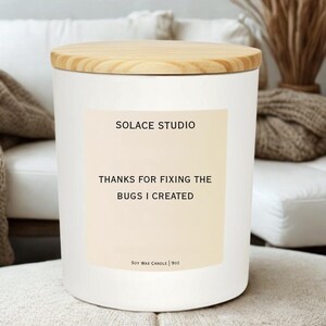 May include: A white soy wax candle with a wooden lid. The candle has a label with the text "SOLACE STUDIO" and "THANKS FOR FIXING THE BUGS I CREATED". The candle is 9 oz.