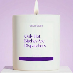 May include: A white soy wax candle with a lit wick. The candle has a purple label with the text "Solace Studio" and "Only Hot Bitches Are Dispatchers." The candle is 9oz.