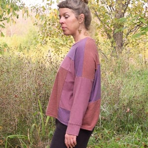 May include: A long-sleeved patchwork top in shades of purple and brown. The top has a round neckline and a relaxed fit. The person is wearing black trousers. The background is a natural outdoor setting with trees and tall grass.