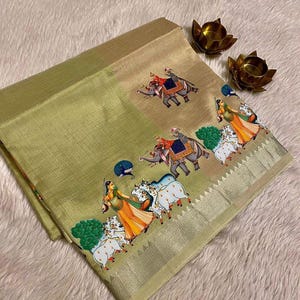 May include: A folded, light green textile with a detailed border. The textile features a repeating pattern of elephants and figures in traditional attire. The border is a metallic gold color. Two small, gold-colored lotus flower candle holders are in the background.