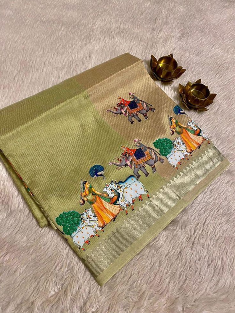 May include: A folded, light green textile with a detailed border. The textile features a repeating pattern of elephants and figures in traditional attire. The border is a metallic gold color. Two small, gold-colored lotus flower candle holders are in the background.