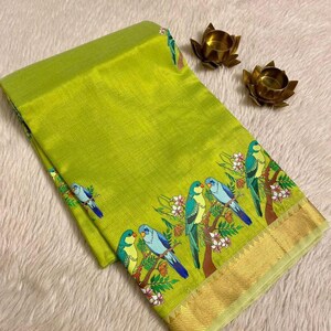 May include: A lime green textile with a detailed border featuring colorful parrots perched on flowering branches. The fabric has a subtle sheen, and the border is accented with a gold trim. Two small, gold lotus-shaped candle holders are also visible.