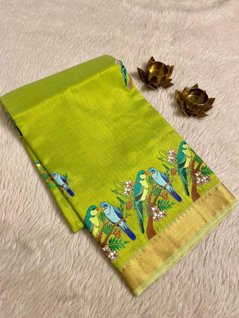 May include: A lime green textile with a detailed border featuring colorful parrots perched on flowering branches. The fabric has a subtle sheen, and the border is accented with a gold trim. Two small, gold lotus-shaped candle holders are also visible.
