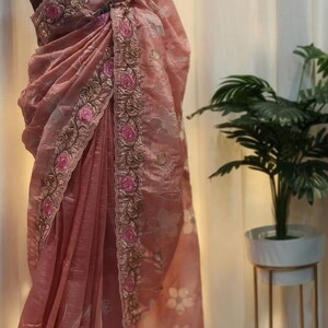 Peut inclure: Un sari rose poudr&eacute; avec des broderies florales complexes. Le sari pr&eacute;sente une bordure d&eacute;taill&eacute;e avec des roses roses et des accents dor&eacute;s. Le tissu se drape &eacute;l&eacute;gamment, mettant en valeur les motifs floraux d&eacute;licats.