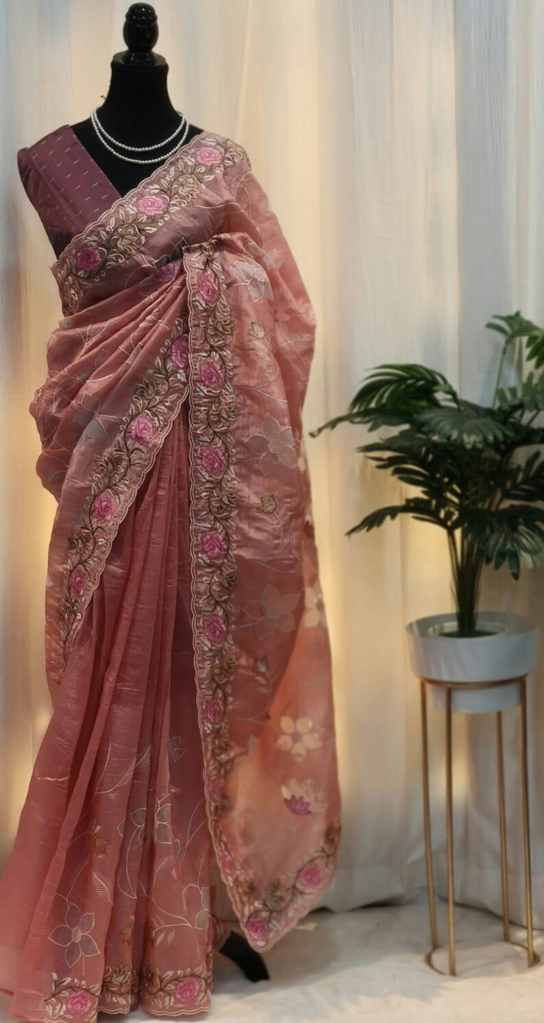 Peut inclure: Un sari rose poudr&eacute; avec des broderies florales complexes. Le sari pr&eacute;sente une bordure d&eacute;taill&eacute;e avec des roses roses et des accents dor&eacute;s. Le tissu se drape &eacute;l&eacute;gamment, mettant en valeur les motifs floraux d&eacute;licats.