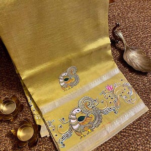 May include: Mustard yellow sari with intricate peacock embroidery. The sari features a detailed border with peacock and floral designs, accented with silver trim. The fabric has a subtle ribbed texture, and the overall aesthetic is elegant and traditional.