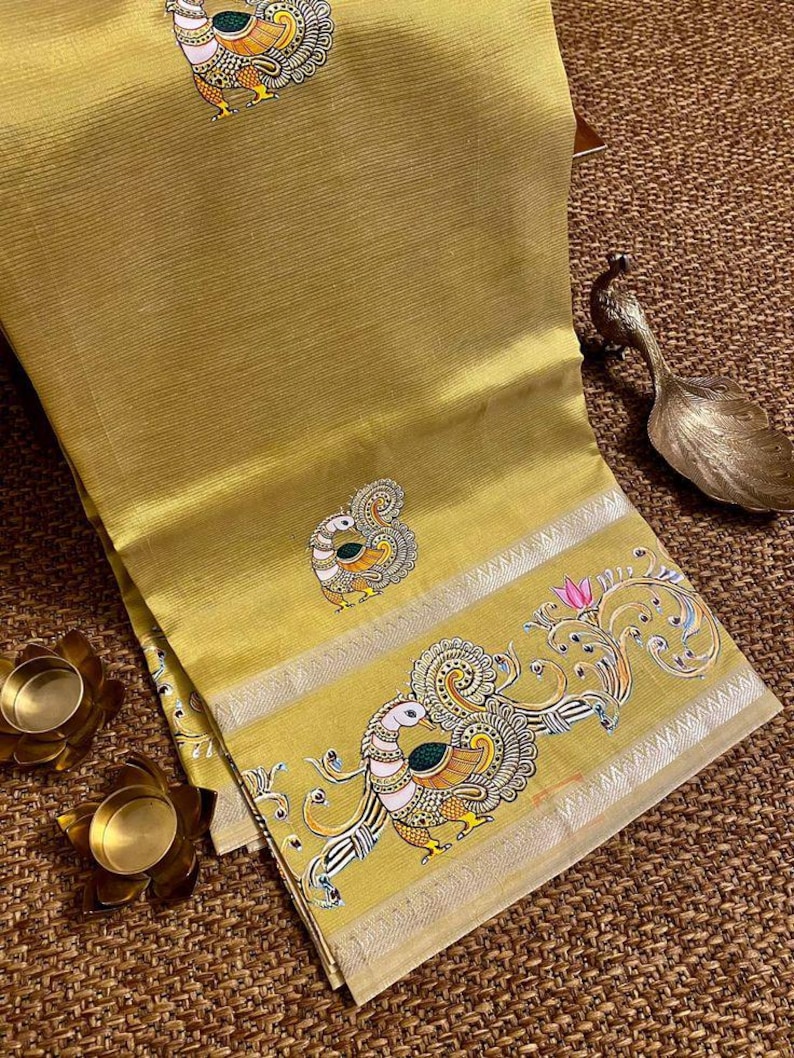 May include: Mustard yellow sari with intricate peacock embroidery. The sari features a detailed border with peacock and floral designs, accented with silver trim. The fabric has a subtle ribbed texture, and the overall aesthetic is elegant and traditional.