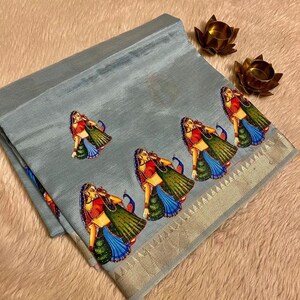 May include: A light blue sari with a decorative border and repeating illustrations of women in traditional attire. The sari features a detailed design with vibrant colors and a metallic gold border.