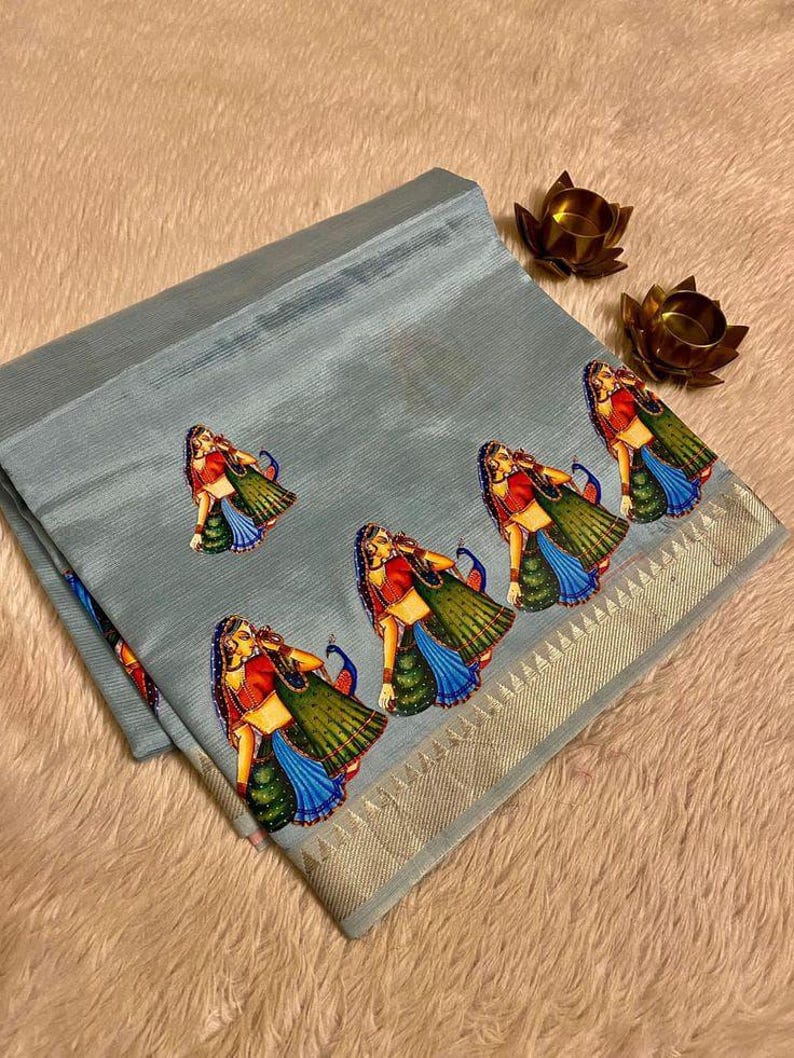 May include: A light blue sari with a decorative border and repeating illustrations of women in traditional attire. The sari features a detailed design with vibrant colors and a metallic gold border.