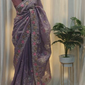 Peut inclure: Un sari lavande avec broderie florale. Le sari est drap&eacute; sur un mannequin, mettant en valeur la bordure florale d&eacute;taill&eacute;e avec des accents roses et dor&eacute;s. Le tissu semble transparent et l&eacute;ger, avec un &eacute;clat subtil.