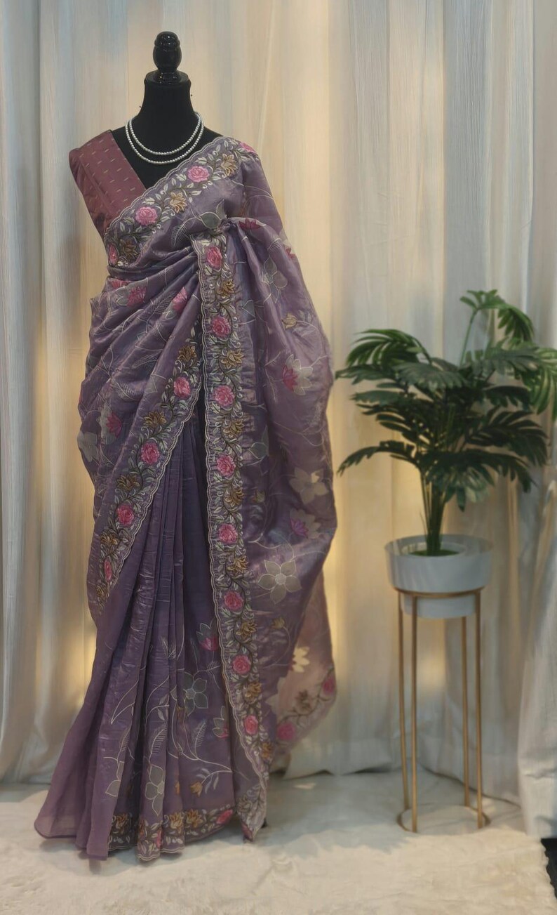 Peut inclure: Un sari lavande avec broderie florale. Le sari est drap&eacute; sur un mannequin, mettant en valeur la bordure florale d&eacute;taill&eacute;e avec des accents roses et dor&eacute;s. Le tissu semble transparent et l&eacute;ger, avec un &eacute;clat subtil.