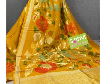 May include: Mustard yellow sari with floral patterns in orange, green, and gold. The sari features a gold border and the text "Sanvi". The fabric appears to be lightweight and flowing, suitable for special occasions.