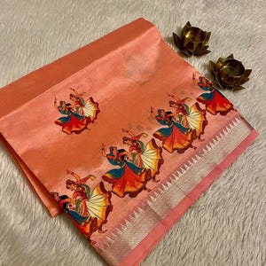 May include: Peach-colored fabric with a repeating pattern of dancing figures in colorful attire. The fabric has a silver border and is folded, showcasing the design. Two small, gold-colored lotus flower candle holders are in the background.