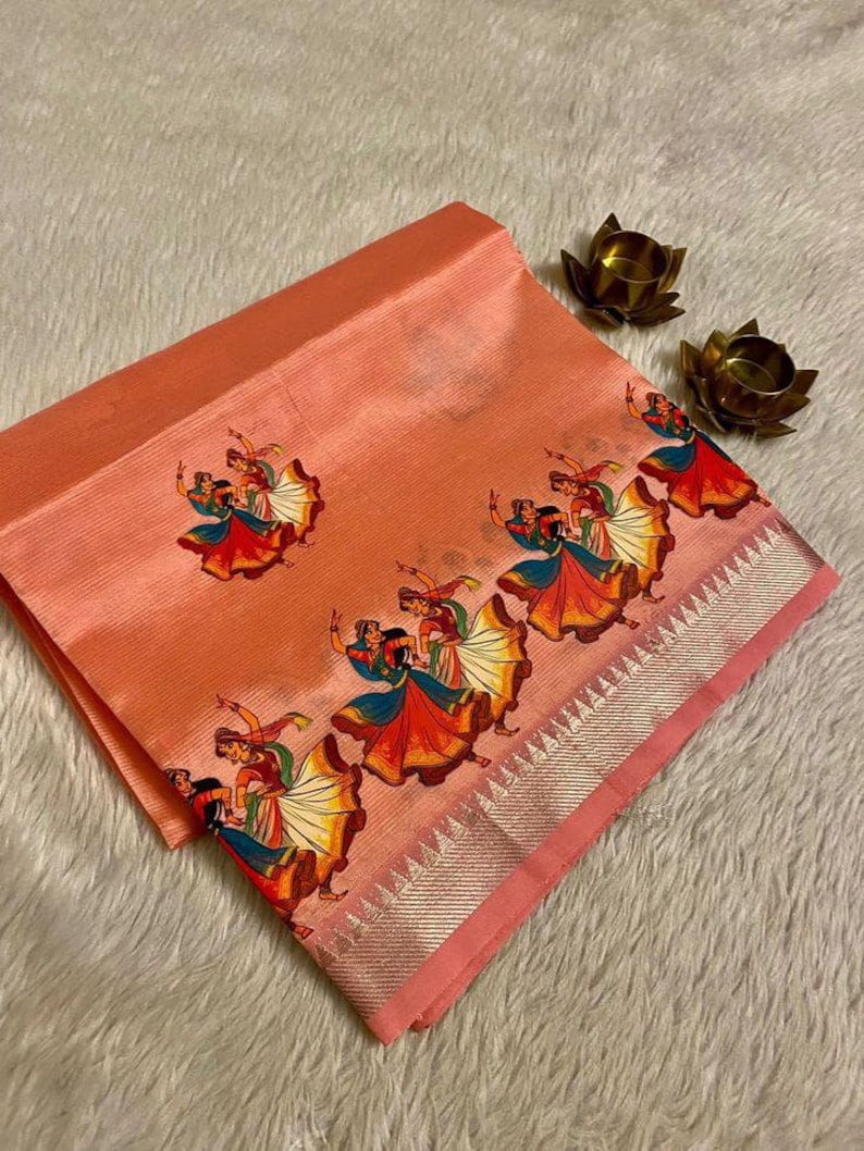 May include: Peach-colored fabric with a repeating pattern of dancing figures in colorful attire. The fabric has a silver border and is folded, showcasing the design. Two small, gold-colored lotus flower candle holders are in the background.