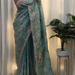 Peut inclure: Un sari vert d'eau avec des broderies florales complexes en rose, or et blanc. Le sari est drap&eacute; sur un mannequin, mettant en valeur son tissu d&eacute;licat et transparent. Le design pr&eacute;sente une bordure d&eacute;taill&eacute;e et des motifs floraux.