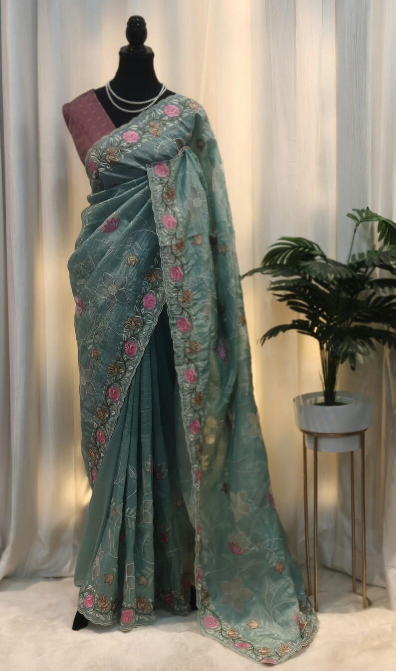 Peut inclure: Un sari vert d'eau avec des broderies florales complexes en rose, or et blanc. Le sari est drap&eacute; sur un mannequin, mettant en valeur son tissu d&eacute;licat et transparent. Le design pr&eacute;sente une bordure d&eacute;taill&eacute;e et des motifs floraux.