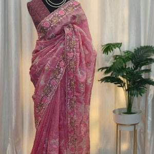 Peut inclure: Un sari rose avec des broderies florales et un chemisier assorti est drap&eacute; sur un mannequin. Le sari pr&eacute;sente des broderies d&eacute;taill&eacute;es le long des bordures et des motifs floraux. Un collier de perles orne le cou du mannequin.