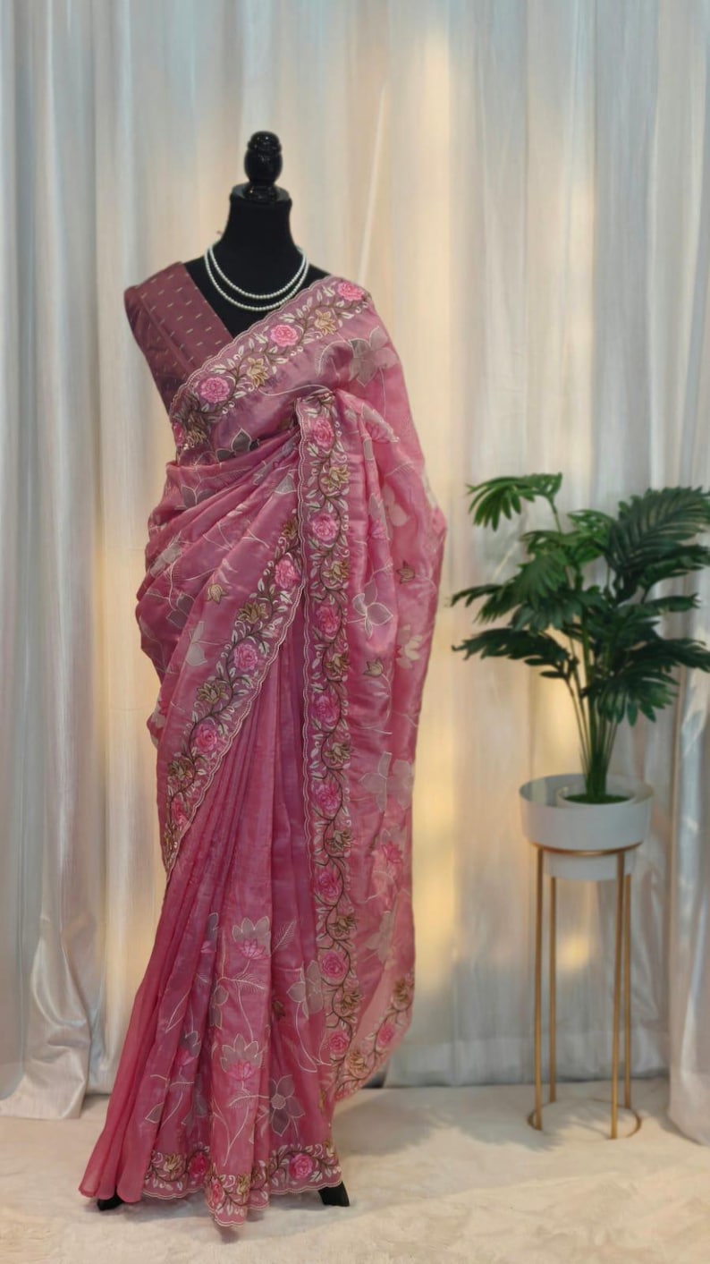 Peut inclure: Un sari rose avec des broderies florales et un chemisier assorti est drap&eacute; sur un mannequin. Le sari pr&eacute;sente des broderies d&eacute;taill&eacute;es le long des bordures et des motifs floraux. Un collier de perles orne le cou du mannequin.