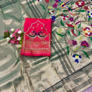 Puede incluir: Un sari verde salvia con brillo metálico, decorado con un patrón de enredadera floral colorida y bordes de rayas verde oscuro. Una pieza de tela rosa brillante doblada con bordado de pavo real dorado y detalles florales descansa encima.