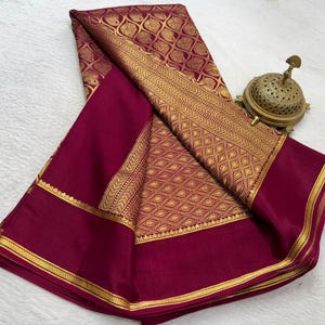 May include: A deep magenta saree featuring a prominent gold zari border and an intricate gold paisley pattern woven throughout the fabric. The textile displays a rich, solid magenta section contrasted by the metallic gold detailing and motifs.