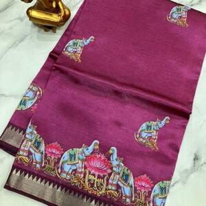 May include: A magenta-colored textile with an elephant and lotus flower pattern. The border features a silver and dark purple design. The elephants are light blue with decorative accents. The textile is folded and draped.