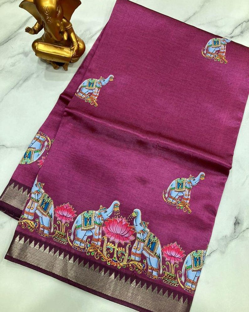 May include: A magenta-colored textile with an elephant and lotus flower pattern. The border features a silver and dark purple design. The elephants are light blue with decorative accents. The textile is folded and draped.