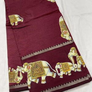 May include: A maroon textile with a repeating pattern of white elephants adorned with gold and yellow decorative elements. The fabric has a subtle striped texture and a decorative border.