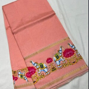 May include: A peach-colored textile with a decorative border featuring elephants, lotus flowers, and ornate gold accents. The fabric is folded, showcasing the detailed design along the edges.