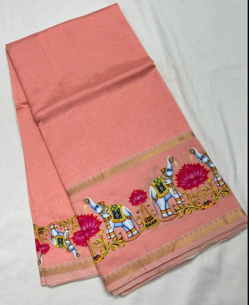 May include: A peach-colored textile with a decorative border featuring elephants, lotus flowers, and ornate gold accents. The fabric is folded, showcasing the detailed design along the edges.